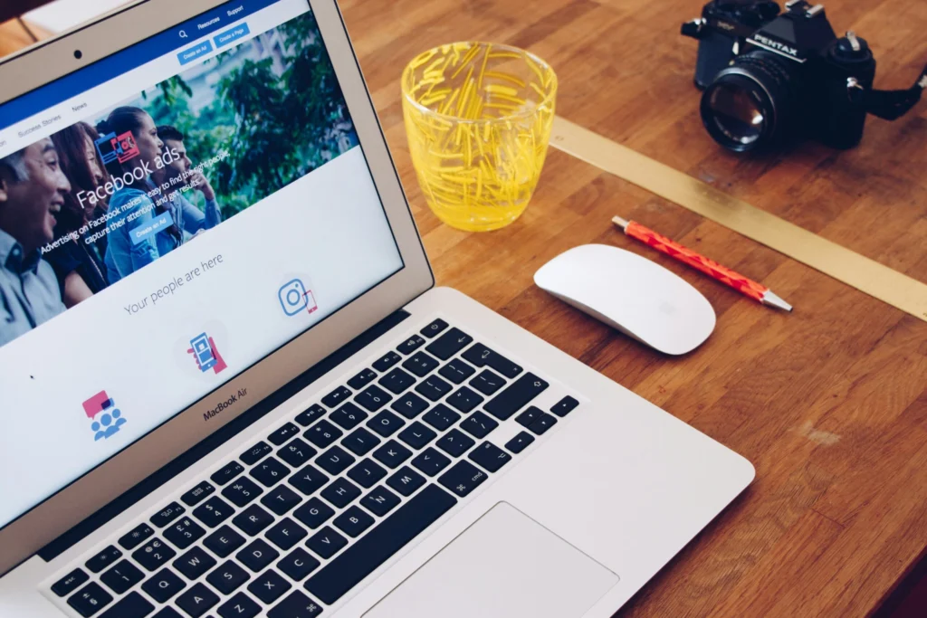 Laptop com a página de anúncios do Facebook aberta, acompanhado de uma câmera, um copo amarelo e uma caneta em uma mesa de madeira.